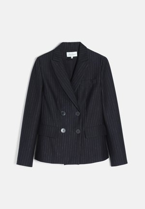 Femme portant un costume noir à fines rayures et double boutonnage, tenant un sac à main noir avec une bandoulière chaîne dorée.