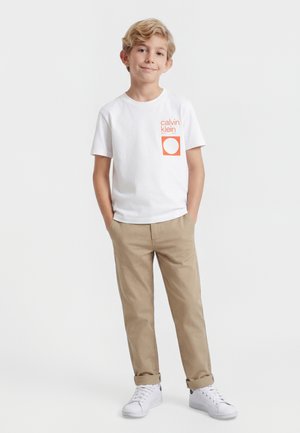 Garçon debout les mains dans les poches, portant un t-shirt blanc Calvin Klein, un pantalon beige et des baskets blanches sur un fond uni.