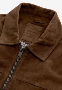 Chaqueta de ante marrón con cierre de cremallera, cuello puntiagudo y etiqueta de cuero con marca. El forro interior presenta un patrón geométrico. Textura suave.
