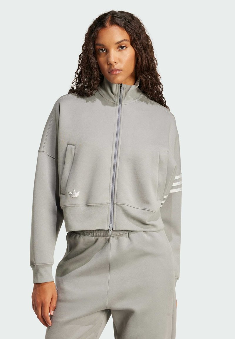 Chaqueta gris cortada con cremallera, hecha de una tela suave, con un alto cuello, dos bolsillos frontales y tres franjas blancas en las mangas.