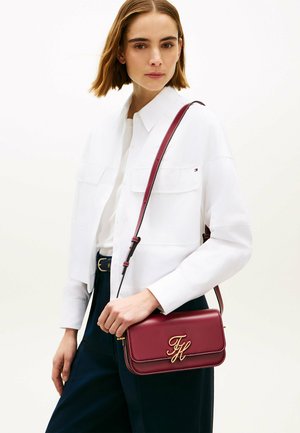 Kvinde iført en hvid skjorte med knapper og marineblå bukser, bærende en burgundy crossbody taske med guldinitialer på den forreste klap.