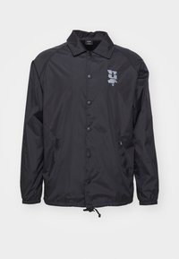 Musta tuulitakki, joka on valmistettu sileästä nailonista, jossa on napitettava etuosa, kiristettävä helmavarsi ja hopeinen logo vasemmassa rinnassa.