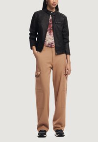 Giacca nera in finta pelle con colletto alto, abbinata a pantaloni cargo beige con grandi tasche e una vestibilità comoda. Sneakers nere completano il look.