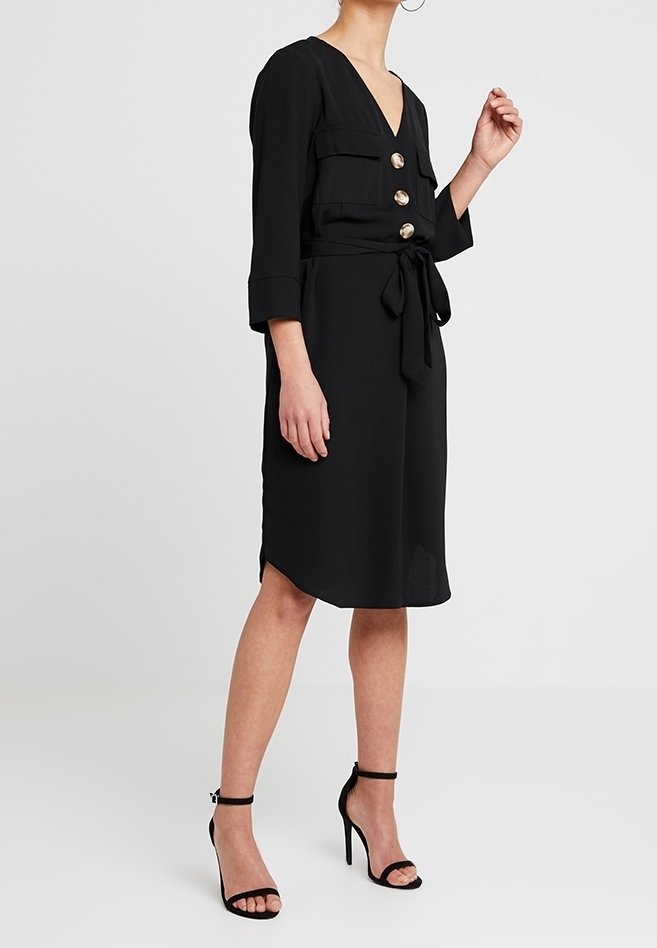 Robe noire avec un décolleté en V profond, des manches trois-quarts et des poches avant. Présente de grands boutons dorés et une ceinture à nouer à la taille. Tissu lisse.