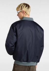 Marineblauer Bomberjacke mit glattem, glänzendem Außenmaterial, gerippten Bündchen und einem hellblauen Kragen. Verfügt über eine Tasche am linken Arm.