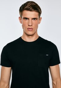 Jaunas vyras trumpais rudais plaukais, dėvintis paprastą juodą marškinėlį be apykaklės, atsisukęs į priekį su neutraliu veido išraiška šviesiame fone.