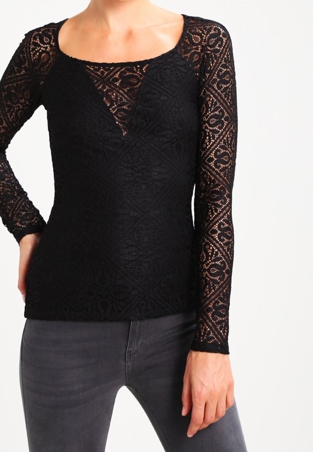 Personne portant un haut noir en dentelle à manches longues avec des motifs géométriques et un jean gris foncé, main gauche posée sur la hanche, visible des épaules aux hanches.