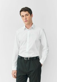 Weißes Langarmhemd mit spitzem Kragen und Knopfleiste, kombiniert mit schwarzen Hosen und einem schwarzen Gürtel mit silberner Schnalle.