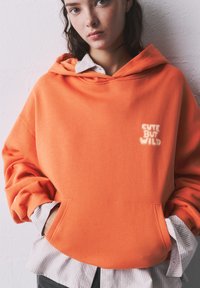 Felpa arancione con tasca anteriore e testo "CUTE BUT WILD". Indossata sopra una maglietta a righe grigie e bianche. Tessuto morbido, vestibilità rilassata.