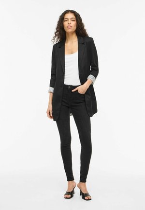 Frau mit lockigem Haar, die einen schwarzen Blazer über einem weißen Top, schwarze Skinny-Jeans und schwarze offene Sandaletten trägt, stehend mit einer Hand in der Tasche.