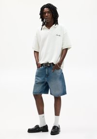 Polo shirt bianco a maniche corte con una texture sottile e logo nero, abbinato a pantaloni corti in denim blu, scarpe nere e calze bianche.