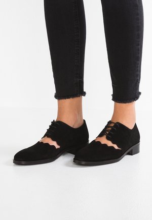 Chaussures en suède noir avec des bords festonnés et des découpages, dotées d'un laçage et d'un petit talon carré. Associées à un jean noir effiloché.