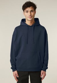 Hoodie bleu marine en tissu doux, avec une grande poche avant, des cordons de serrage et des poignets côtelés, porté sur un pantalon noir.