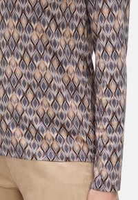 Nahaufnahme eines Langarmoberteils mit einem geometrischen Muster aus braunen, beigefarbenen und grauen Rauten, getragen zu hellbraunen Hosen.