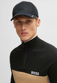 Schwarze Cap mit tonal gestaltetem "BOSS"-Logo, kombiniert mit einem Sweatshirt mit Reißverschluss, das schwarze und beige Farbkombinationen, strukturierte Strickoptik und taillierte Passform aufweist.