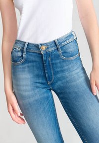 Jean en denim bleu à coupe ajustée, taille haute et décoloration subtile. Présente des boutons dorés et des détails de couture sur les poches.