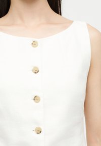 Femme portant un haut blanc sans manches avec un col carré et quatre boutons dorés sur le devant.
