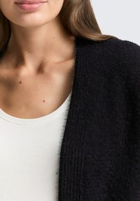 Schwarze, strukturierte Strickjacke mit einem weichen, flauschigen Aussehen, getragen über einem weißen Rundhalsoberteil. Nahaufnahme des Hals- und Schulterbereichs.