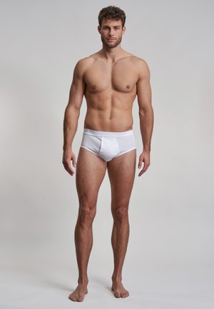 Weiße Baumwollunterhose mit einer engen Passform, ausgestattet mit einem weichen Bund und flachen Nähten für zusätzlichen Komfort. Das Modell steht barfuß vor einem neutralen Hintergrund.