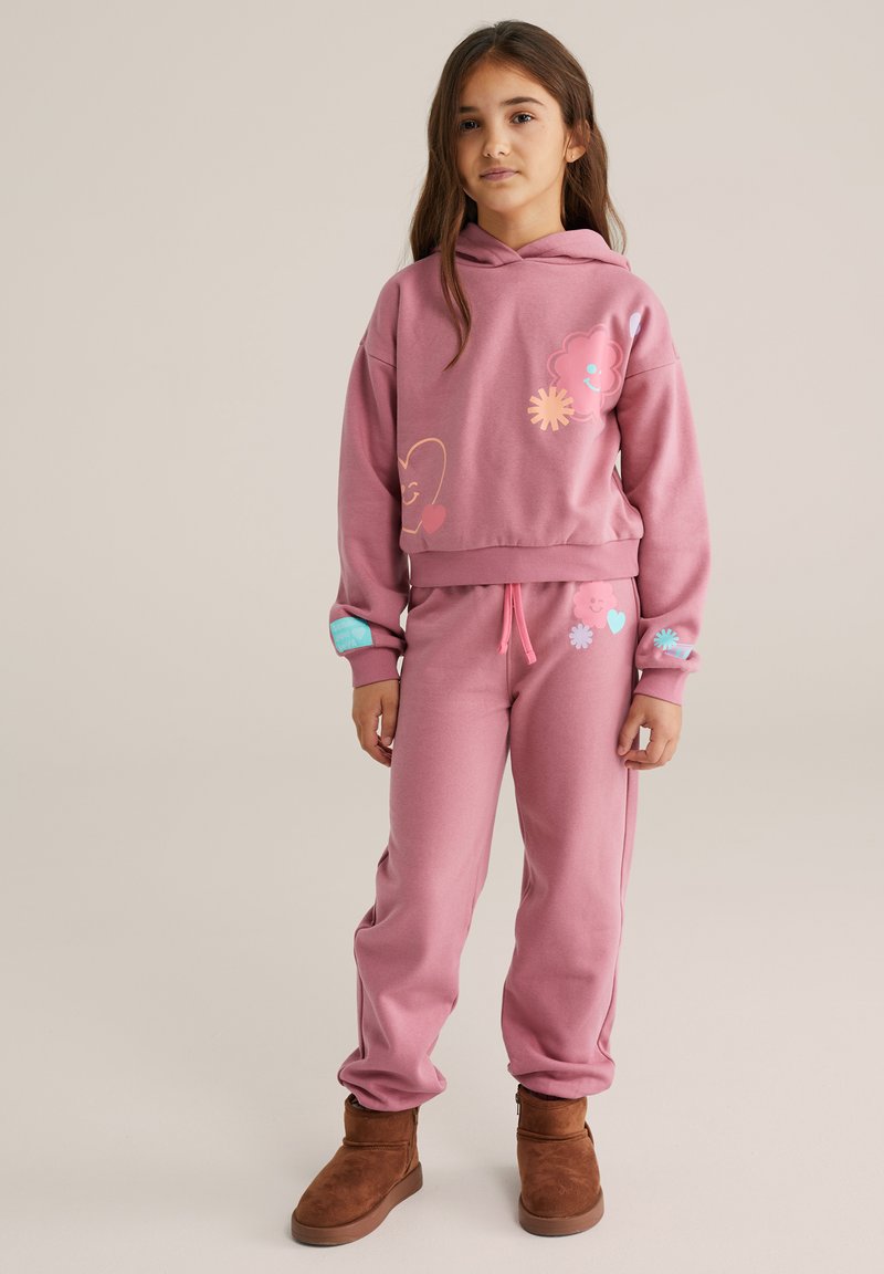 Lyserød hættetrøje og matchende joggingbukser lavet af blødt stof, med farverige blomster- og smiley-grafikker. Brune ankelstøvler fuldender outfittet.