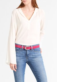 Weiße Langarmbluse mit V-Ausschnitt, kombiniert mit blauen Jeans und einem leuchtend pinken Gürtel mit einer silbernen Schnalle.