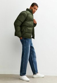 En mörkgrön puffjacka med hög krage, quiltad struktur och dragkedjefickor, i kombination med lösa blå jeans och vita sneakers.