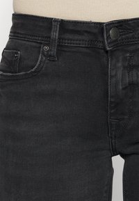 Pantalones vaqueros de mezclilla negra con un ajuste slim, que presentan bolsillos frontales, un cierre de botón y costuras en contraste. Textura suave con un ligero brillo.