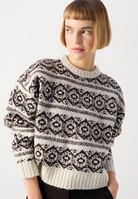 Maglione crema con intricatati motivi geometrici marroni, orlo e polsini a costine e scollatura rotonda. Tessuto lavorato a maglia e vestibilità rilassata.