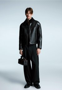 Crna kožna jakna s umetnom krznom na ovratniku, zatvaranje na patentni zatvarač i glatka površina. Kombinira se s labavim crnim hlačama i malom torbicom.