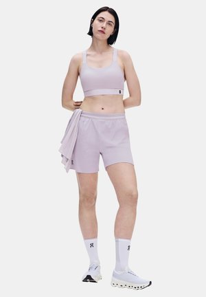 Frau steht mit den Händen hinter dem Rücken, trägt einen hellvioletten Sport-BH, passende Shorts, hält ein Hemd, weiße Socken und weiße Turnschuhe.