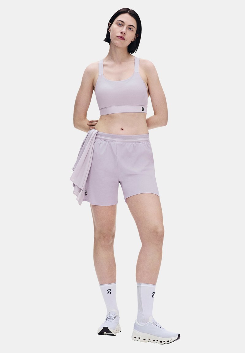 Femme debout, les mains derrière le dos, portant un soutien-gorge de sport violet clair, un short assorti, tenant une chemise, des chaussettes blanches et des baskets blanches.