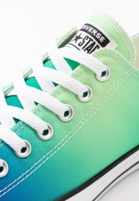 Green and blue gradient canvas sneaker with white laces, silver eyelets, and a black and white logo patch on the tongue.