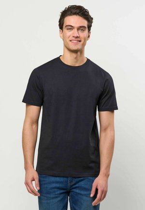 Noor mees tume juustega, kes kannab tavalist musta lühikeste varrukatega T-särki ja siniseid teksapükse, seisab heleda tausta ees ja naeratab.