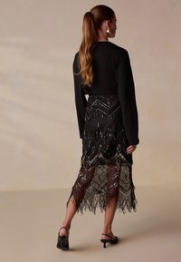 Black long-sleeve top worn with a beaded black and metallic fringed skirt featuring a geometric pattern, knee-length with a flowing hem.