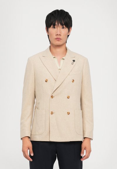 Blazer beige croisé en tissu texturé, avec des boutons marron, deux poches avant et une épingle noire sur le revers.