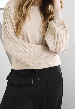 Femme portant un pull côtelé beige avec des manches bouffantes exagérées et un pantalon noir à cordon, posant avec une main près de la tête.