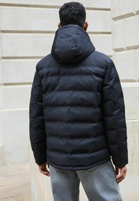 Schwarze Steppjacke mit Kapuze, ausgestattet mit gesteppten Paneelen und einer glatten Textur. Die Rückseite zeigt ein schlankes Design und elastische Bündchen.