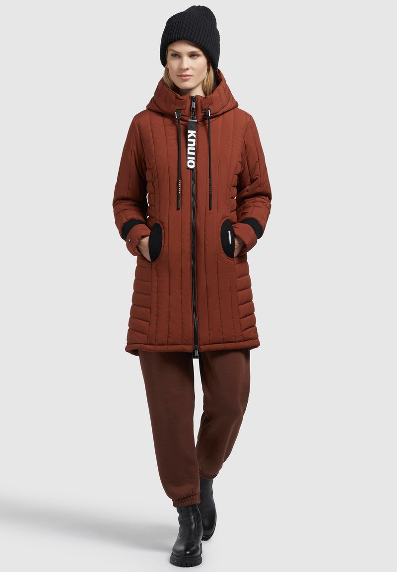 Braune gesteppte Pufferjacke mit schwarzen Akzenten, einer Kordelzugkapuze und seitlichen Taschen, kombiniert mit braunen Hosen und schwarzen Stiefeln.