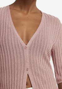 Light pink knit cardigan with a deep V-neck, ribbed texture, short sleeves, and a single button closure at the front.