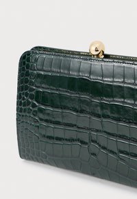 Πράσινο πορτοφόλι με ανάγλυφη επιφάνεια κροκοδείλου με γυαλιστερό φινίρισμα, διαθέτοντας χρυσό κούμπωμα και υφή. Περιλαμβάνει δομημένο σχήμα και ορατά ραψίματα.