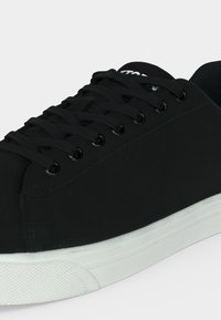 Μαύρο αθλητικό παπούτσι χαμηλής κορυφής με μαύρα κορδόνια και λευκή καουτσούκ σόλα με υφασμένη προστατευτική μύτη για καλύτερη πρόσφυση.