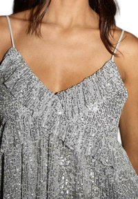 Srebrna bleščeča majica s volanastim ovratnikom, tankimi naramnicami in ohlapnim krojem. Ima sijočo teksturo in večplastno zasnovo.