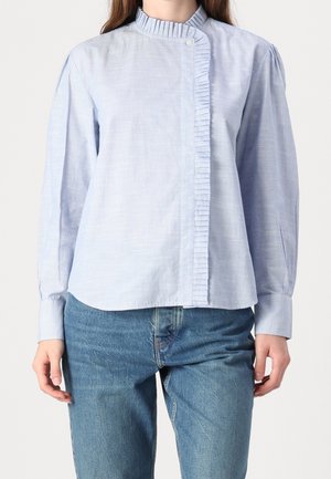 Nainen pukeutunut vaaleansiniseen puseroon, jossa on laskostettu kaulus ja edessä yksityiskohta, yhdistettynä sinisiin farkkuihin, yksiväristä taustaa vasten.