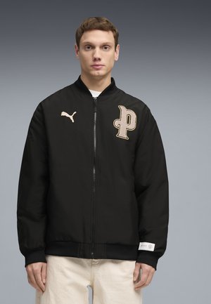 Joven con chaqueta bomber negra de Puma con parche beige en forma de "P" y pantalones color crema sobre fondo gris.