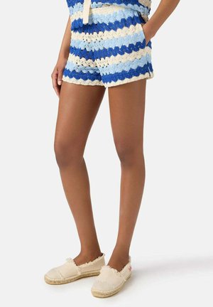 Donna che indossa pantaloncini a righe azzurro e bianco all’uncinetto con tasche e scarpe espadrillas crema con bordi sfrangiati.