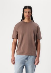 Brown short-sleeve T-shirt with a round neckline, made of cotton, featuring a relaxed fit and straight hem, worn with light blue jeans.