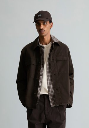 Homme portant une casquette sombre, une veste foncée superposée sur une chemise à carreaux et un t-shirt de couleur claire, debout devant un fond clair uni.