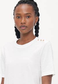Hvid t-shirt lavet af blødt stof, med korte ærmer og rund halsudskæring. Tre dekorative nitter pryder venstre skulder.