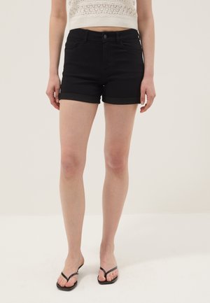 Donna con pantaloncini neri con orlo arrotolato, top corto bianco lavorato a maglia e sandali neri infradito, in piedi contro uno sfondo semplice.