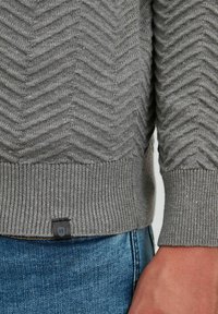 Grå strikket sweater med et fiskebensmønster, ribbede manchetter og et lille sort mærke i kanten. Båret med blå denim jeans.